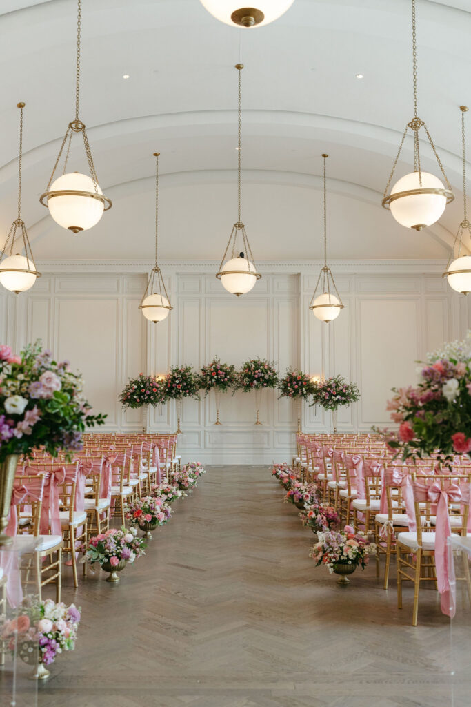 Wedding ceremony space with gold chairs with pink bows tied on the back and vases of pink and purple flowers in the aisle at The Adolphus in Dallas