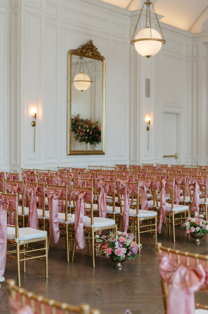 Wedding ceremony space with gold chairs with pink bows tied on the back and vases of pink and purple flowers in the aisle at The Adolphus in Dallas