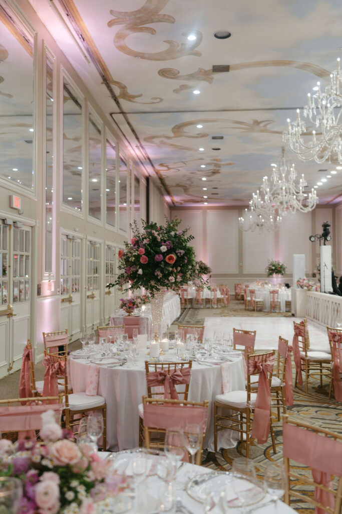 Wedding reception space with gold chairs with pink bows tied on the back around round white tables and vases of pink and purple flowers on top at The Adolphus in Dallas