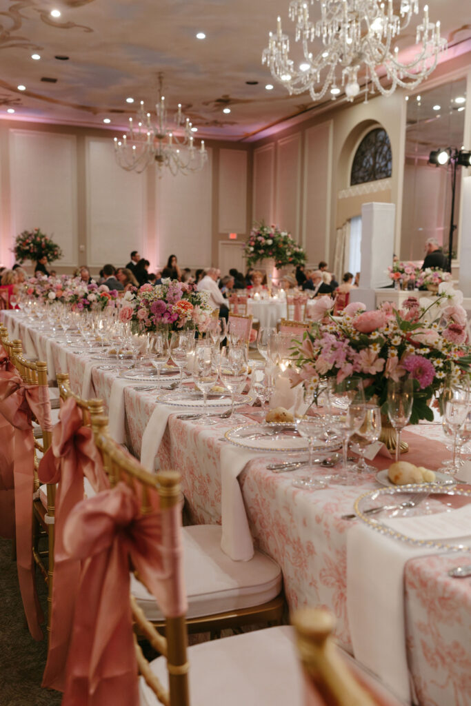 Wedding reception space with gold chairs with pink bows tied on the back by a long white table and vases of pink and purple flowers on top at The Adolphus in Dallas
