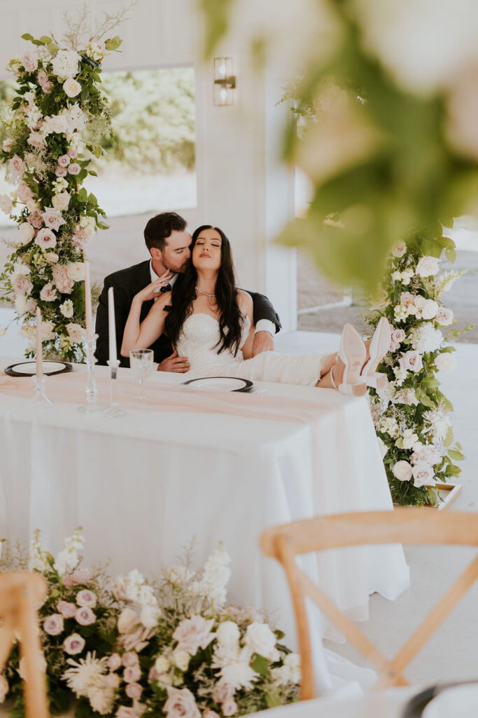 Bride and groom embracing sitting at a white table under a flower arch at a venue in Dallas