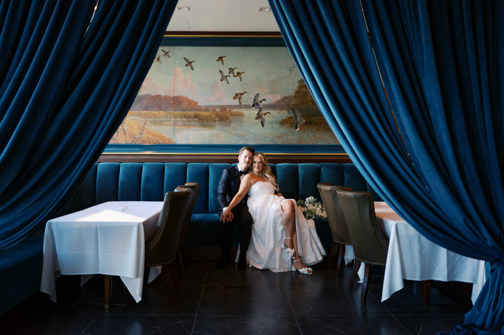 Bride and groom sitting in a restaurant with blue velvet booths and a large painting of flying geese and large blue drapers