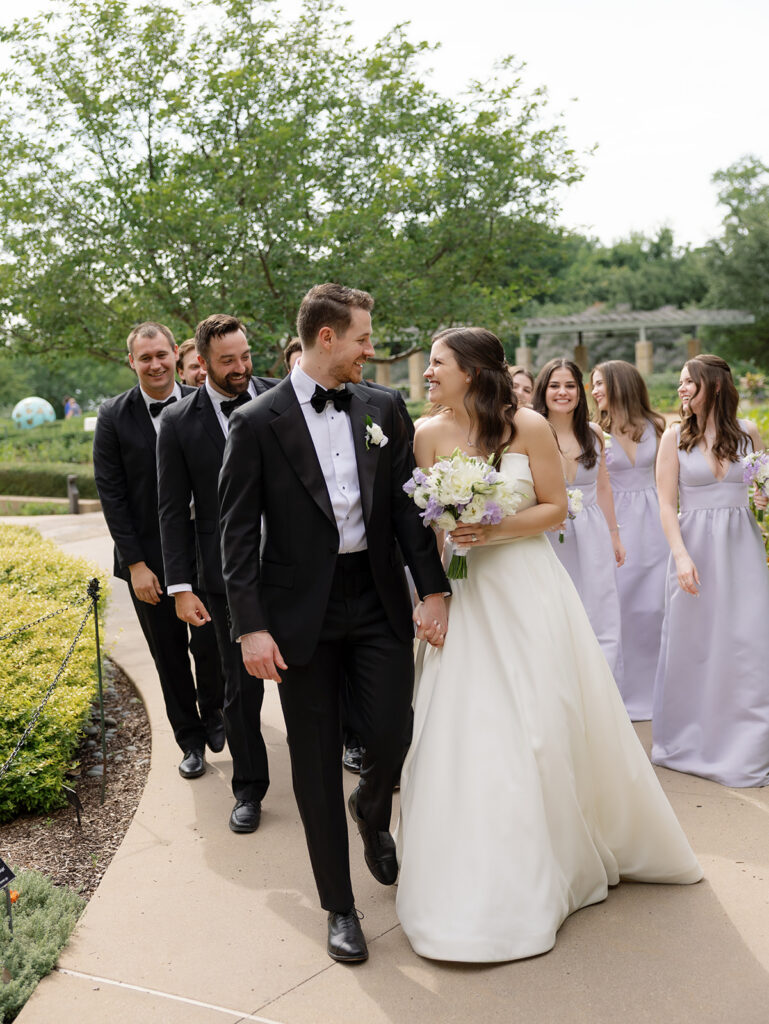 Bride and groom walking outside with groomsmen in black tuxes and bridesmaids in light purple dresses