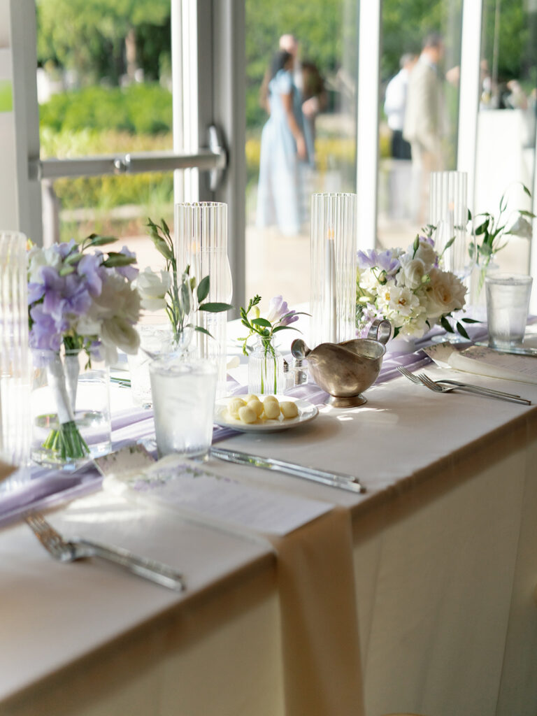 Wedding reception table set with a light purple runner and purple and white flowers in small vases in between tall candles