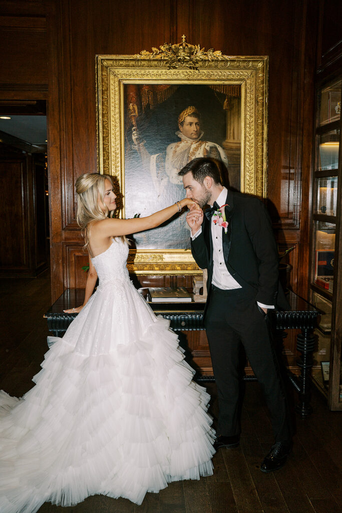 Groom in black tux kissing bride's hand wearing white ballgown while standing in front of a historic oil painting