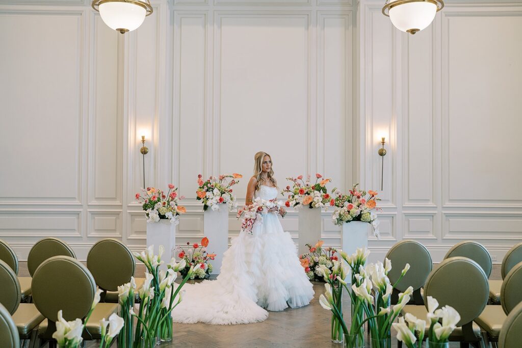 Bride in white ballgown with ruffled skirt standing at front of ceremony space with pink and orange floral arrangements